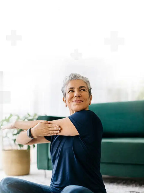 An older woman with short gray hair, wearing a navy blue t-shirt and blue pants, stretching her left arm across her body with her right hand on her left elbow.