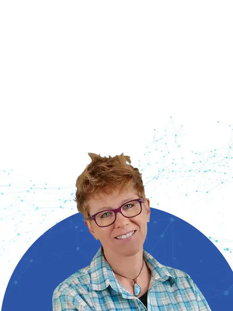 A smiling woman with short, spiky reddish-brown hair and purple-framed glasses, wearing a blue and white plaid shirt and a turquoise pendant.